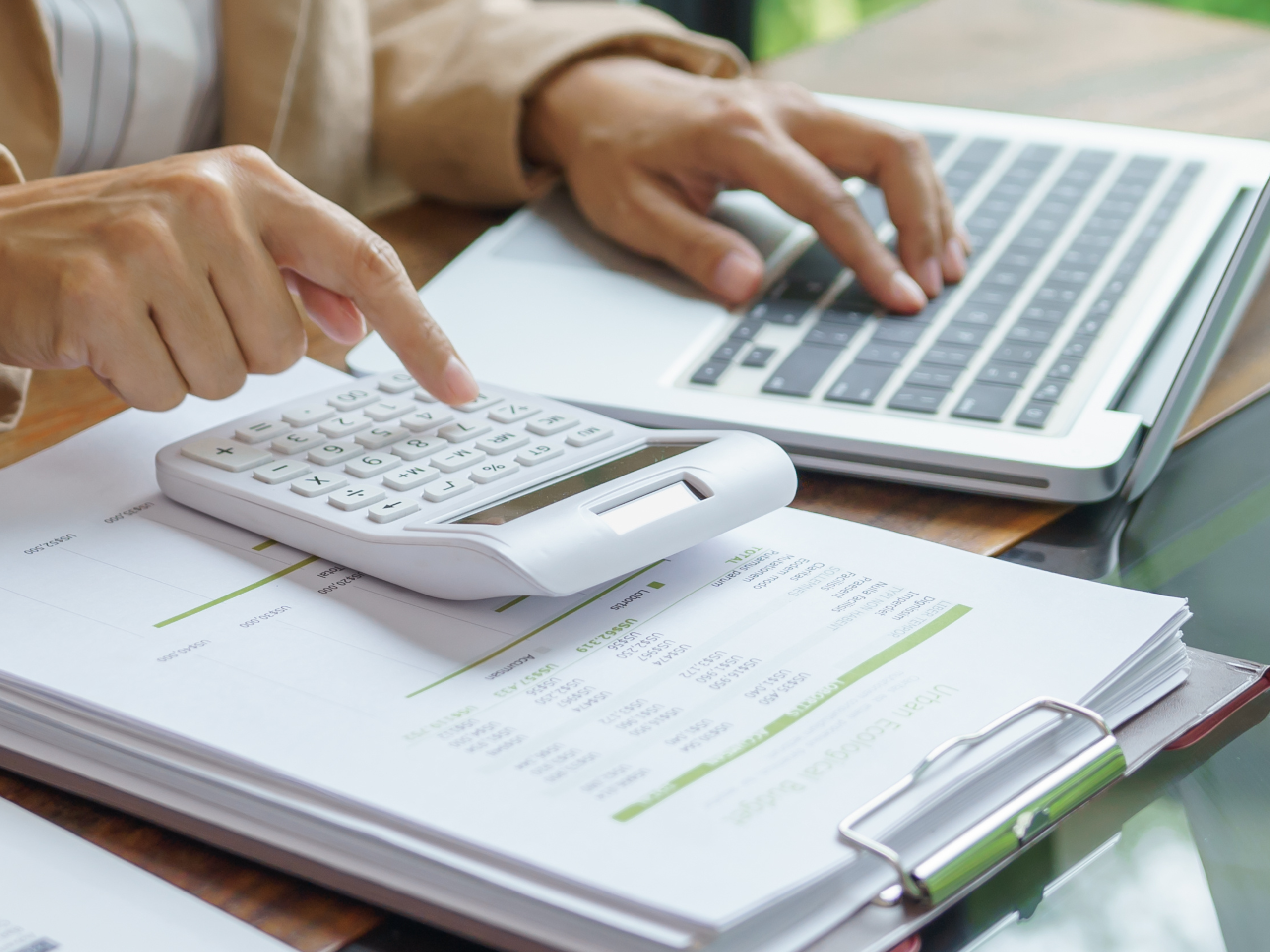 A person using a laptop with a calculator and financial documents, working through financial calculations.